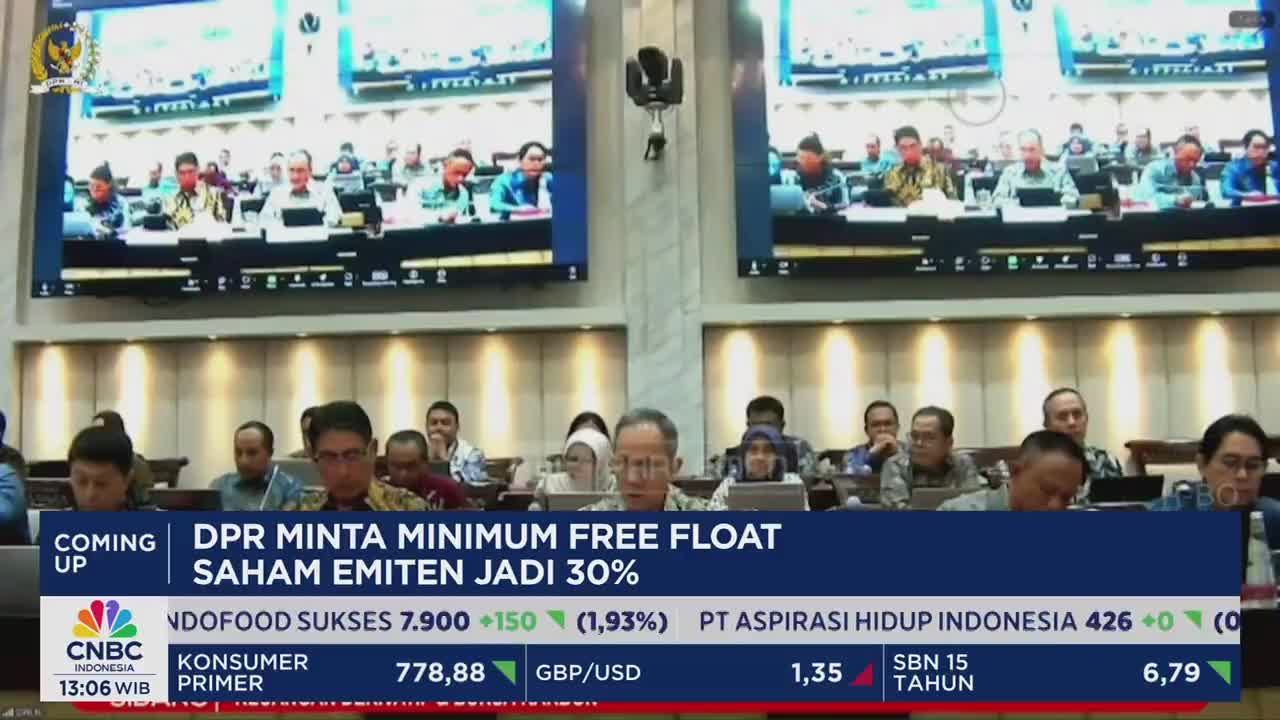 In a large room, people are seated at long tables, some looking at screens displaying a video conference. CNBC Indonesia's news ticker at the bottom of the screen flashes financial information.
