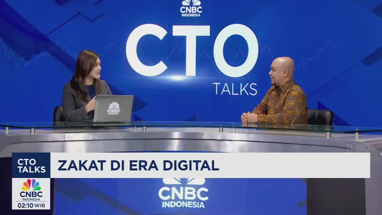 A woman in a grey blazer speaks while gesturing, a laptop open before her. Across the table, a man in a patterned shirt listens intently.