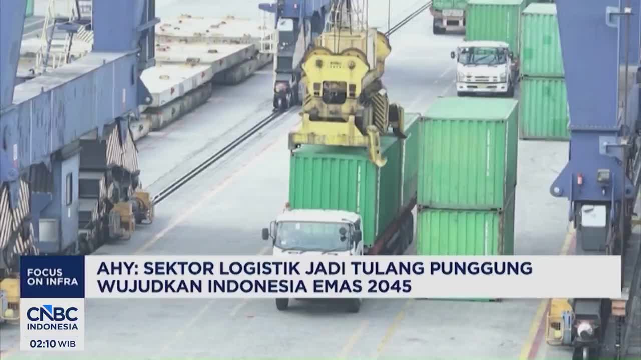 A massive crane lifts a green shipping container, lowering it onto a waiting truck. Nearby, other trucks navigate between stacks of containers at the busy port.