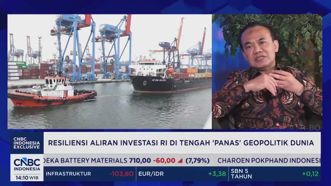 A red and white tugboat maneuvers in the water, guiding a large container ship past towering cranes at the port. On the right, a man in a patterned shirt speaks, his hands gesturing as if explaining something important.