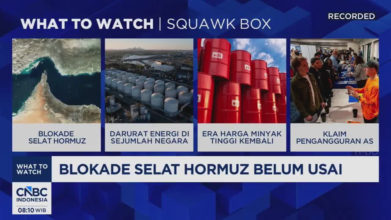 A large body of water, possibly a strait, is bordered by arid land. Nearby, rows of white storage tanks stretch across a landscape under a hazy sky. In the foreground, a stack of bright red oil drums reaches towards a blue sky.