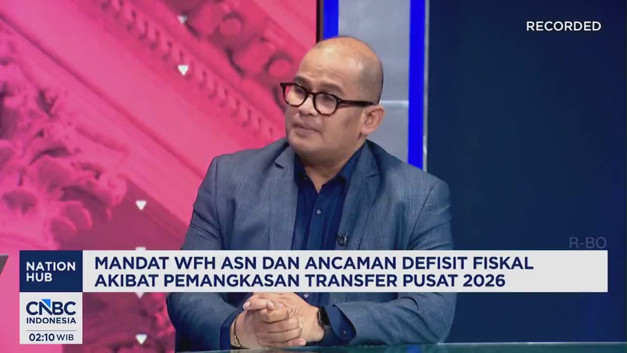 A man in a suit and glasses sits at a table, speaking on CNBC Indonesia. The screen behind him displays text about government mandates and fiscal deficits.