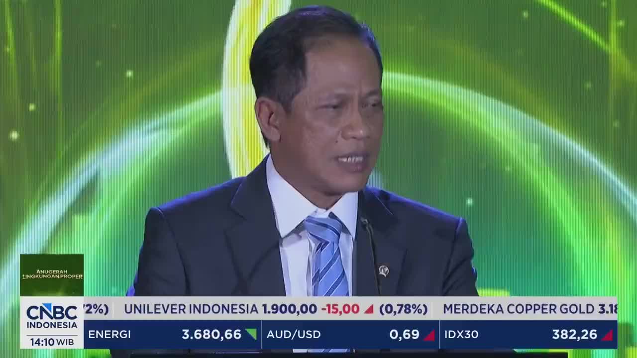A man in a suit speaks, his tie a pattern of blue stripes. Below him, a ticker displays stock information for Unilever Indonesia and other companies. A man in a suit speaks, his tie a pattern of blue stripes. Below him, a ticker displays stock information for Unilever Indonesia and other companies.