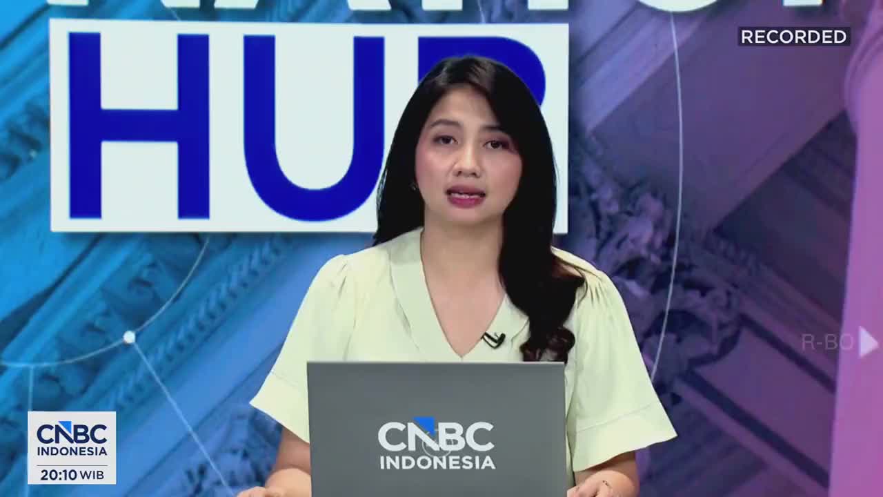 A woman in a pale green blouse speaks directly to the camera, a laptop with the CNBC Indonesia logo in front of her. Behind her, large blue letters spell out "HU," part of a larger graphic.