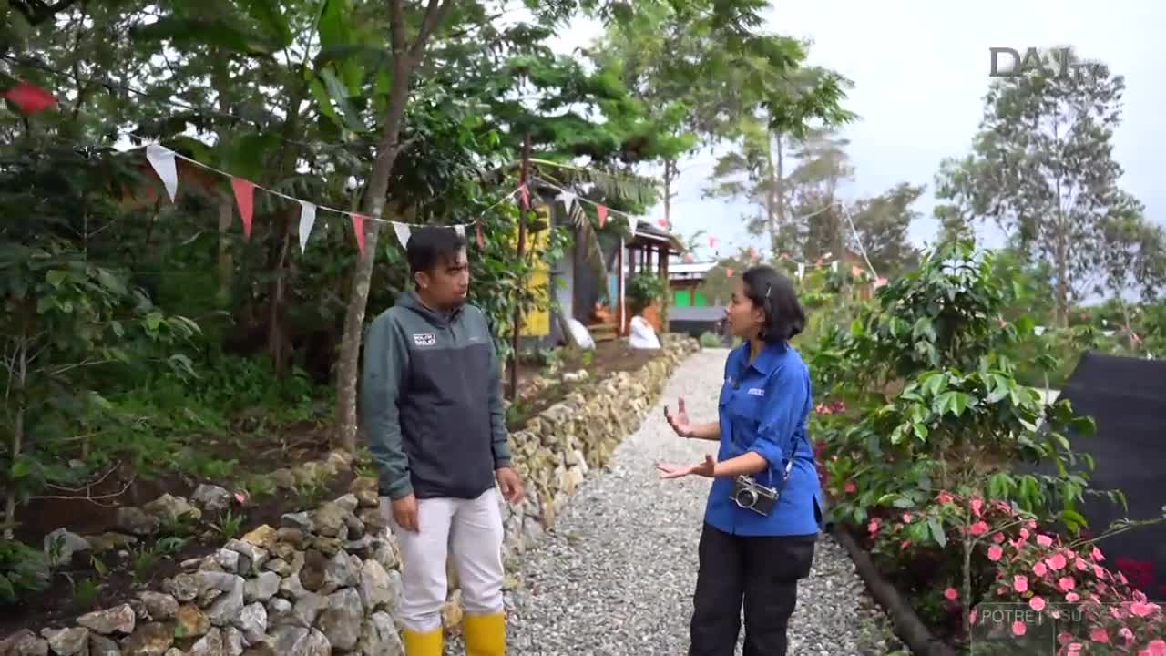 A woman in a blue shirt gestures with her hands, speaking to a man in a jacket and yellow boots. They're on a gravel path, and the DAAI TV logo is visible in the corner.

