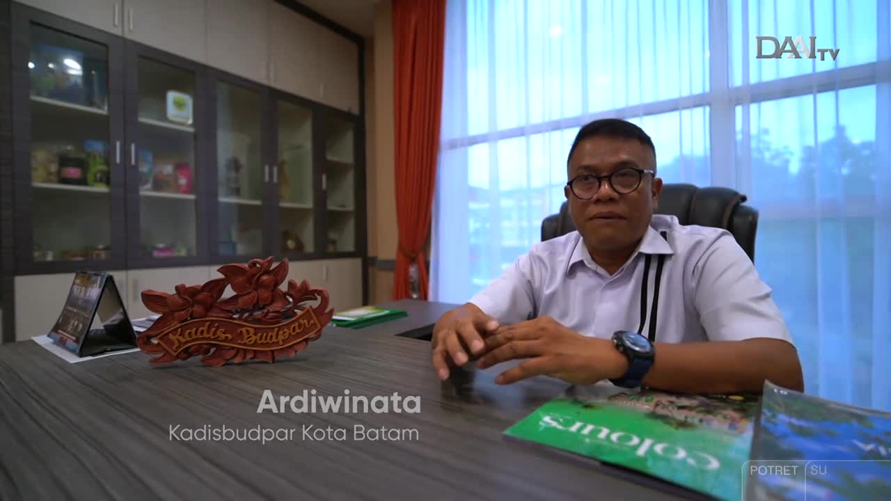 Ardiwinata, Kadisbudpar Kota Batam, sits at his desk, facing the camera for DAAI TV. A wooden carving and some brochures rest on the table in front of him.

