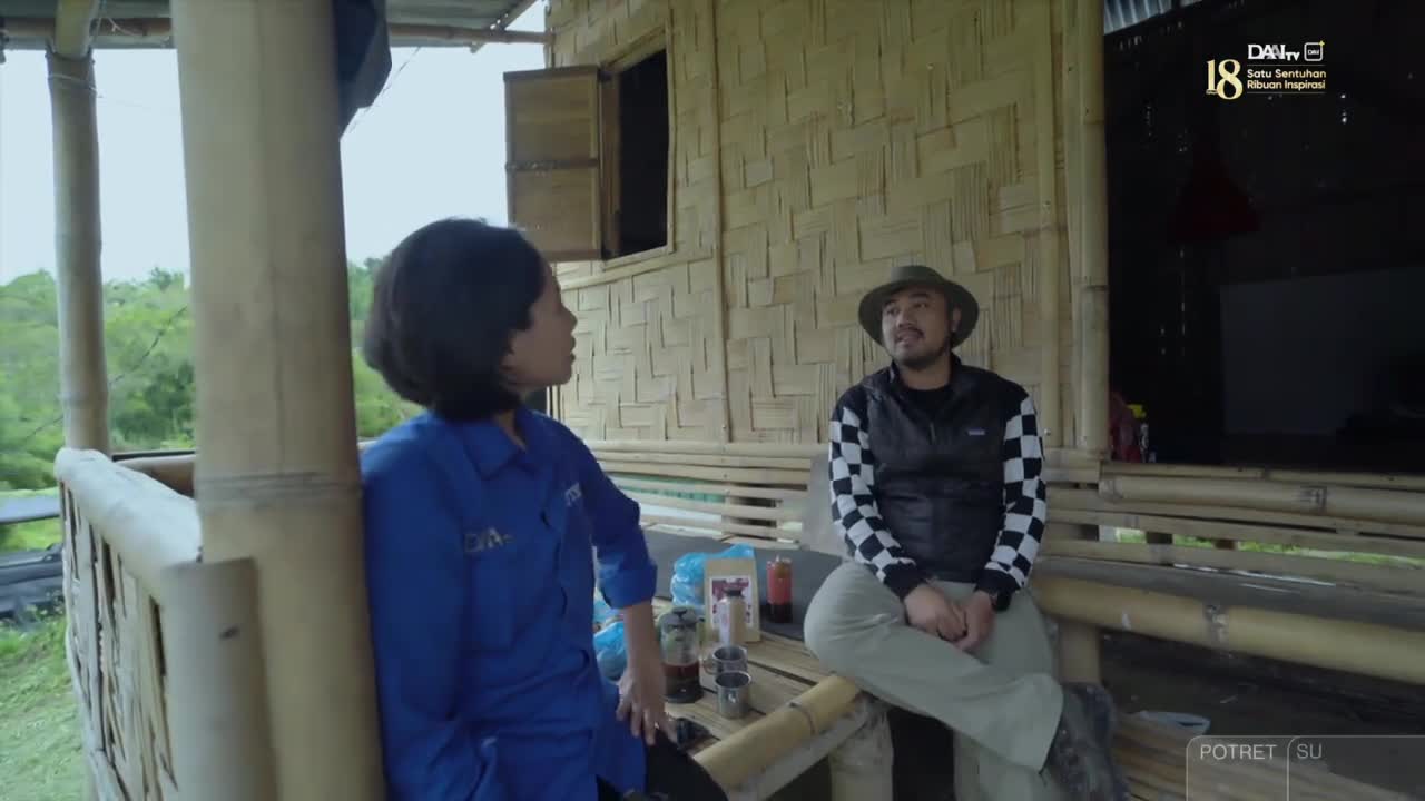 A woman in a blue shirt is talking to a man wearing a hat and a checkered jacket, both seated on a bamboo porch. They're outside, and the DAAI TV logo is visible in the corner.
