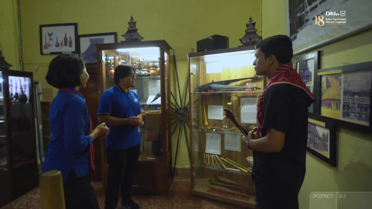Three people stand in a museum, examining artifacts displayed in glass cases. A man in a black shirt holds a long, dark object, possibly a ceremonial weapon, as he speaks.
