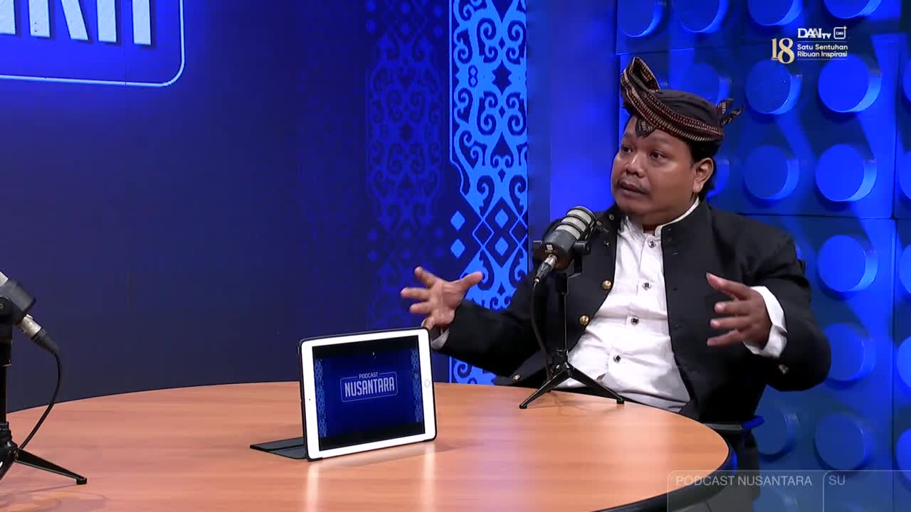 A man in a traditional Indonesian headdress and jacket gestures with both hands, speaking into a microphone.  The set is for DAAI TV's "Podcast Nusantara" program.
