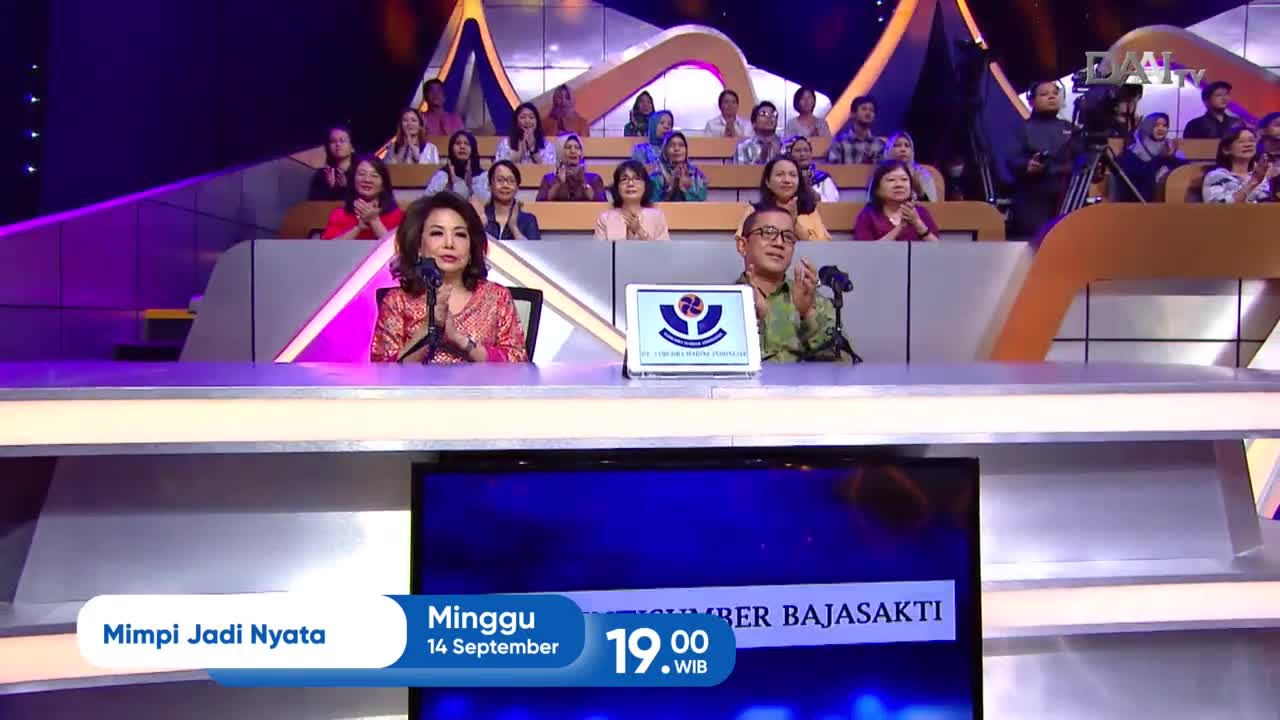 The two hosts, a woman in a patterned red top and a man in a green military-style shirt, are clapping in front of a small screen displaying a logo. Behind them, the audience in the Indonesian DAAI TV studio applauds.
