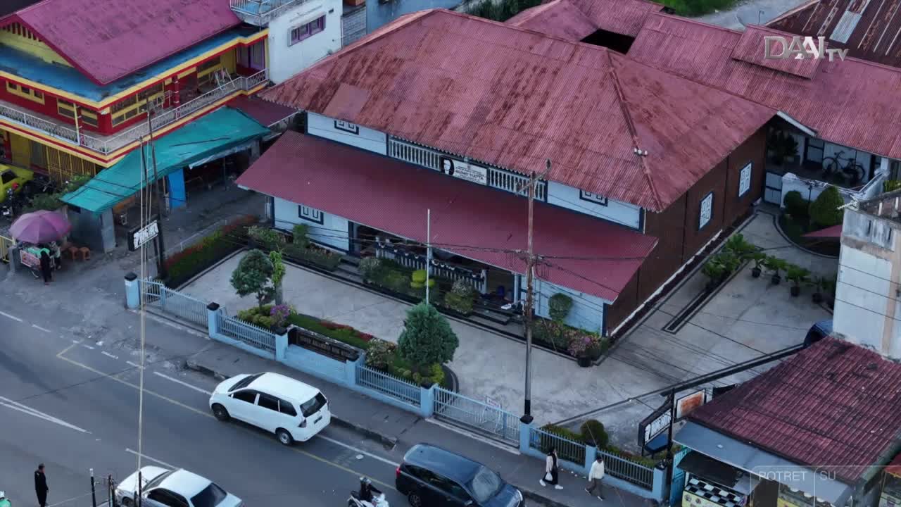 Cars and scooters travel along a street in front of a building with a DAAI TV logo. The building has a red roof and a white facade, and there's a small garden in front.
