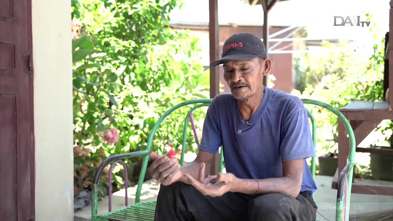 An older man in a blue shirt and a cap sits on a green metal chair, gesturing with his hands as he speaks. The DAAI TV logo is visible, and the setting appears to be a home in Indonesia, with lush greenery in the background.
