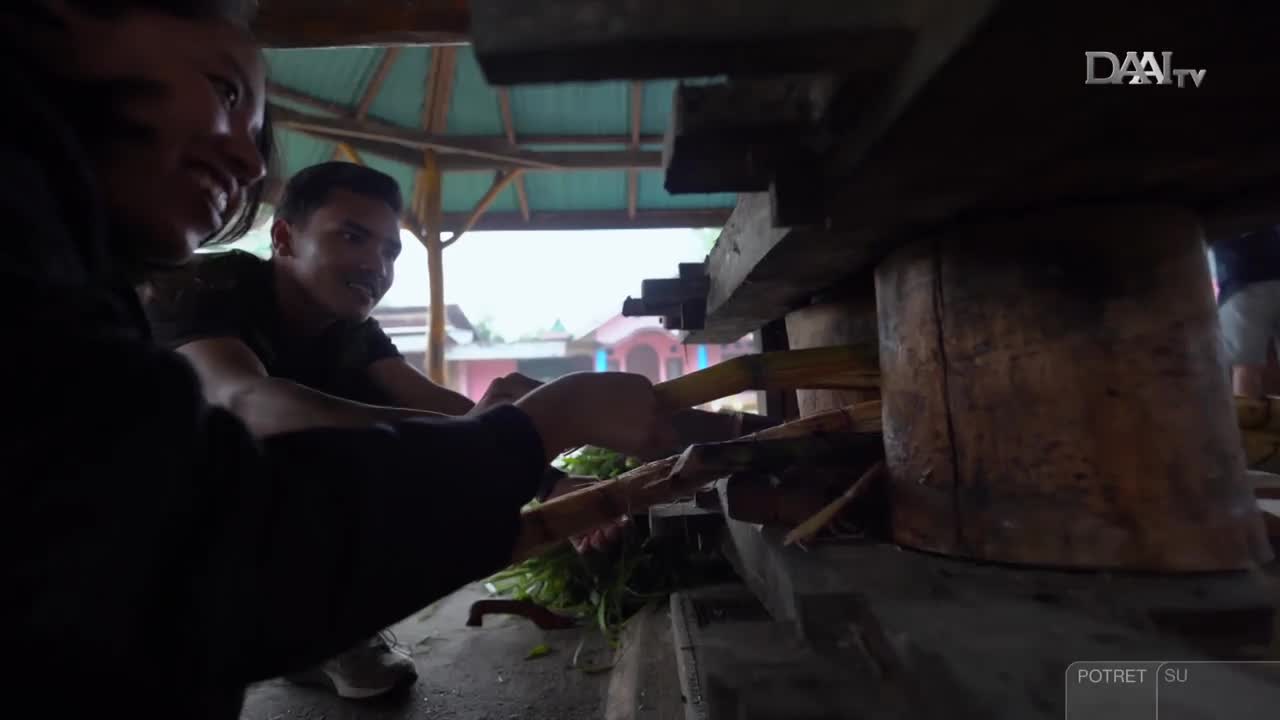 Two people work together, feeding stalks of sugarcane into a large, wooden press. The scene is being filmed by DAAI TV in Indonesia, where the men smile as they work.
