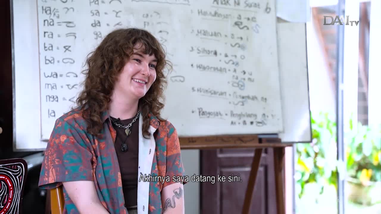 A woman with curly hair smiles, seated in front of a whiteboard covered in Indonesian script. Below her, text appears in Indonesian on the DAAI TV broadcast.

