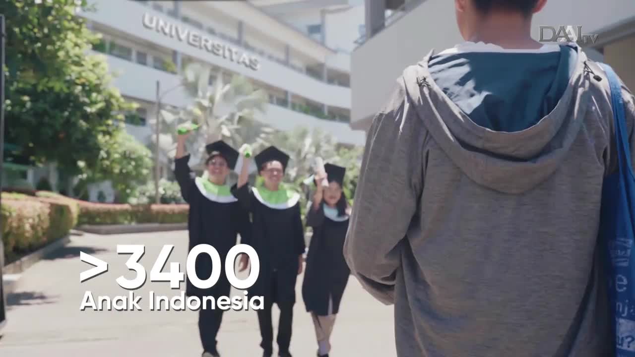 Graduates in black gowns and caps are celebrating, raising their arms in front of a university building. A person in a gray hoodie walks away, partially obscuring the scene, as DAAI TV's logo is visible.
