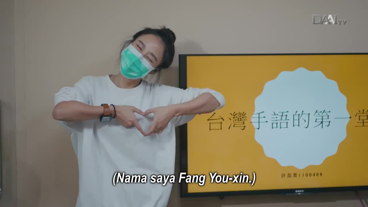 A woman in a mask makes a heart shape with her hands, smiling at the camera. Behind her, a DAAI TV screen displays text in Chinese, likely part of a sign language lesson.
