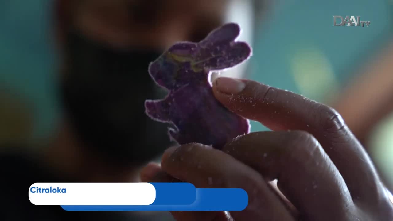 A person holds up a small, purple bunny-shaped object, likely a craft project, for the DAAI TV cameras. The bunny's edges are slightly fuzzy, and the person's fingers are dusted with a fine powder.
