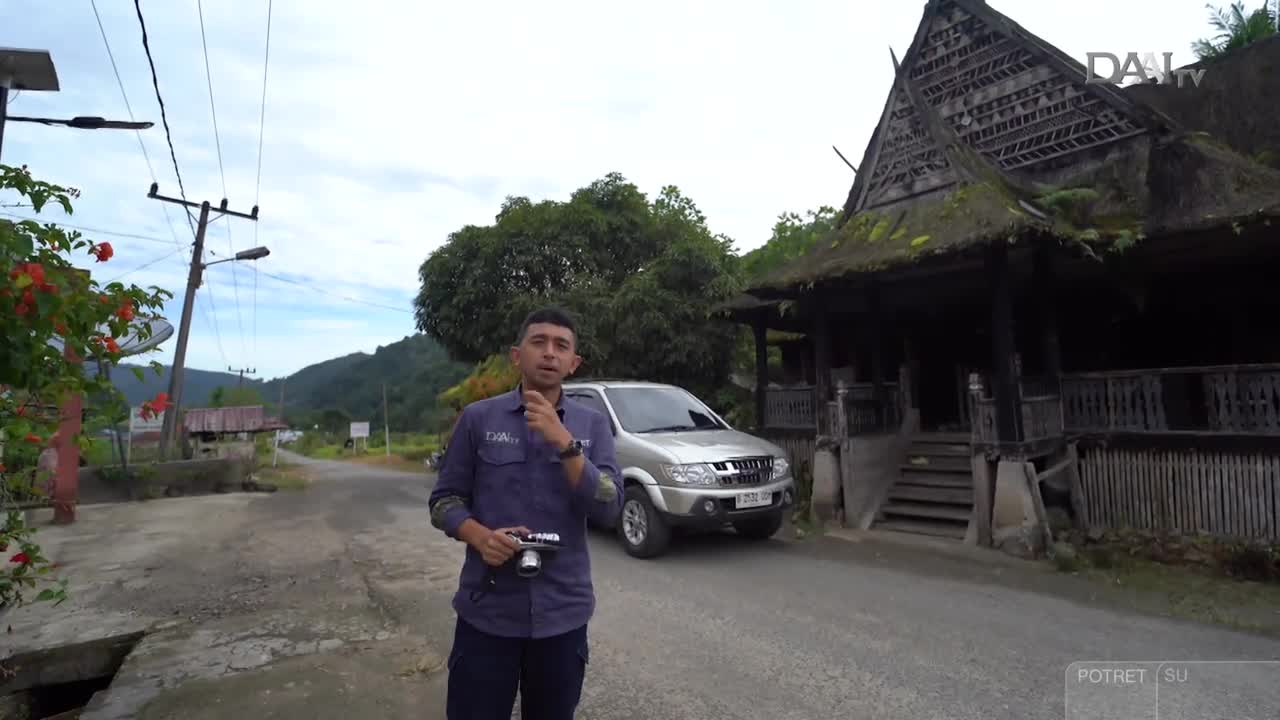 A man, holding a camera, gestures while standing on a road in Indonesia. Behind him, a traditional wooden structure stands next to a silver SUV, with the DAAI TV logo in the corner.
