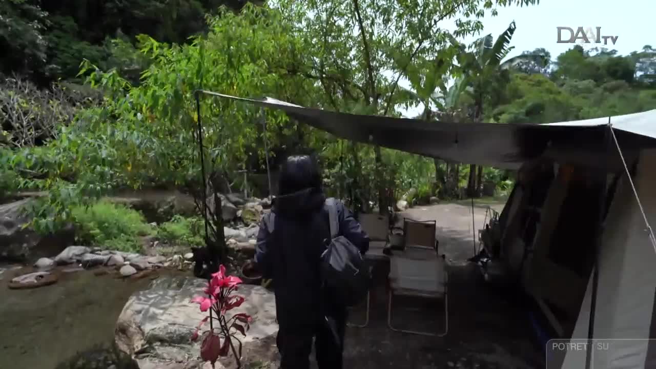 A person walks towards a campsite set up near a stream, carrying a bag over their shoulder. The scene is being filmed, likely for DAAI TV in Indonesia, with a tent and chairs visible in the background.
