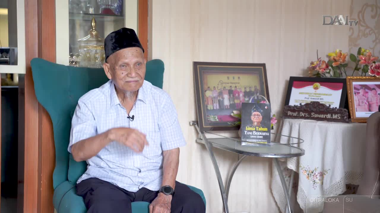 An elderly man in a black cap gestures with his right hand as he speaks, likely during an interview for DAAI TV in Indonesia. Behind him, a table displays a book and framed photos, including one with his name, Prof. Drs. Suwardi, MS.
