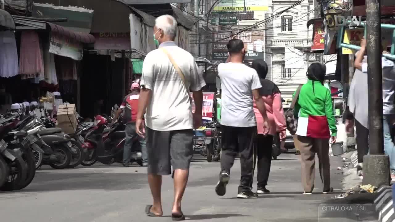 People stroll down a crowded street, some wearing masks, passing shops and parked motorbikes. Overhead, a tangle of wires crisscrosses the sky in this Indonesian market scene.
