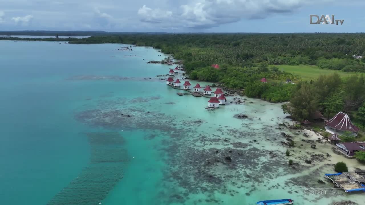 The turquoise water laps gently against the shore, where small houses with red roofs stand on stilts. Lush green trees line the coast, and the sky is overcast.
