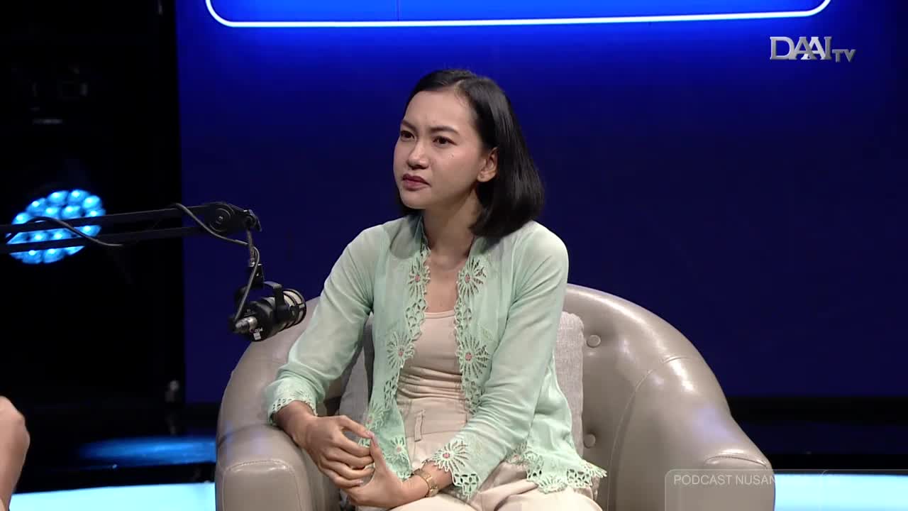 The woman, dressed in a light green kebaya, sits in a plush chair, speaking on a DAAI TV set. A microphone hovers nearby, and a blue light shines in the background.

