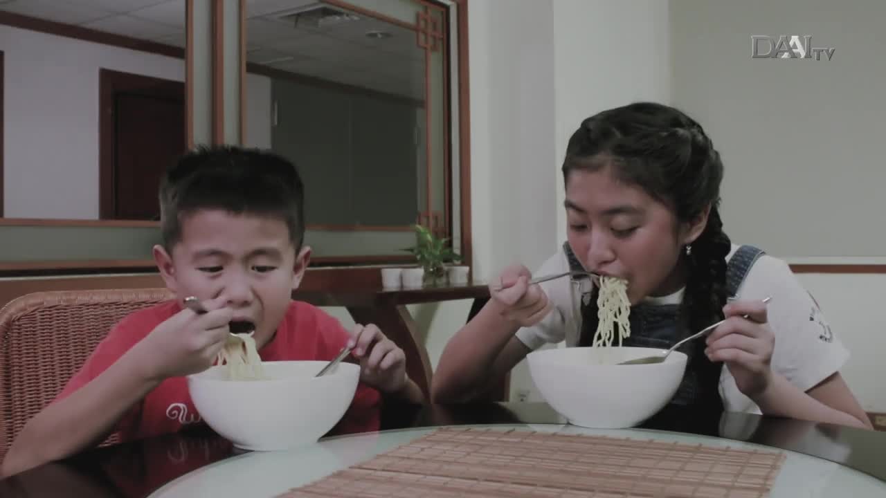 A boy in a red shirt and a girl with braided hair are eating noodles from white bowls. They're using chopsticks and forks, focused on their meal.