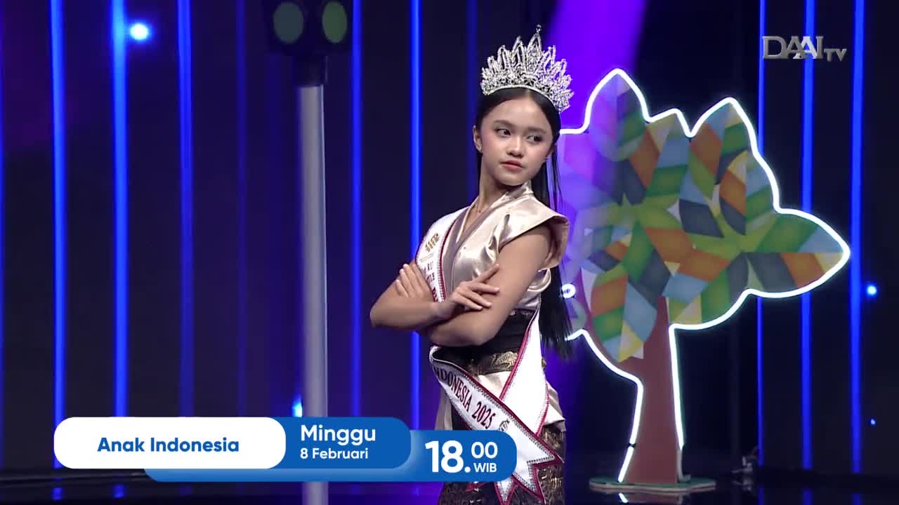 A young woman with a crown stands with her arms crossed, a sash across her chest. She's on a stage with a colorful tree cutout and blue lights behind her, likely for a DAAI TV broadcast in Indonesia.