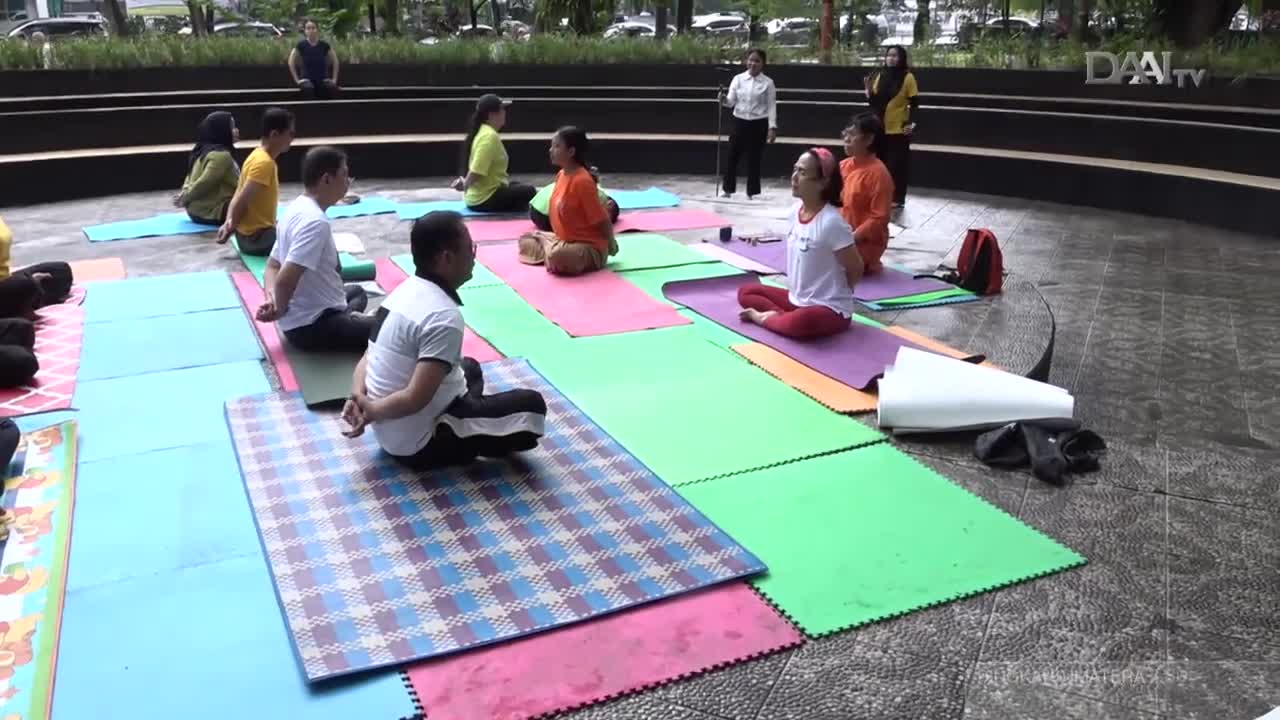 A group of people are sitting cross-legged on colorful mats in an outdoor plaza, their hands clasped behind their backs. Two women in the background, likely instructors for the DAAI TV broadcast, stand observing the session.