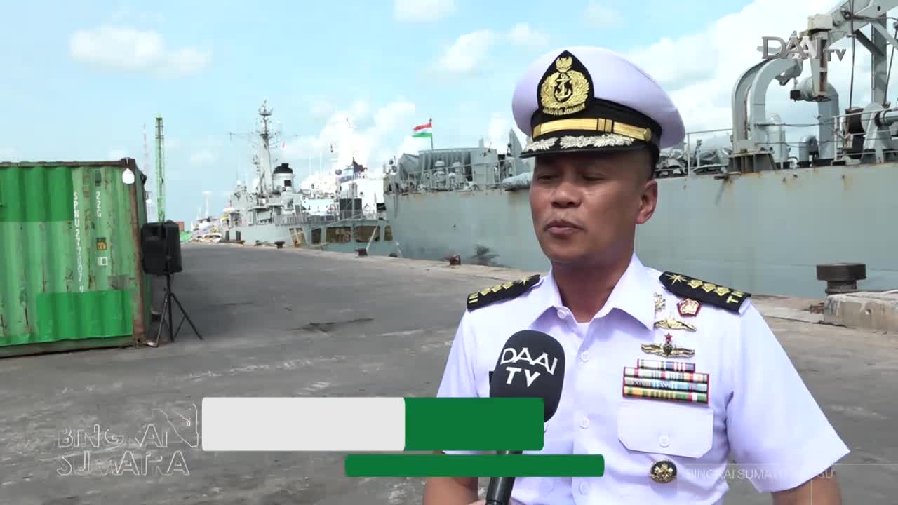 A naval officer speaks into a DAAI TV microphone, his uniform adorned with medals. Behind him, a large ship rests at anchor in the Indonesian harbor.