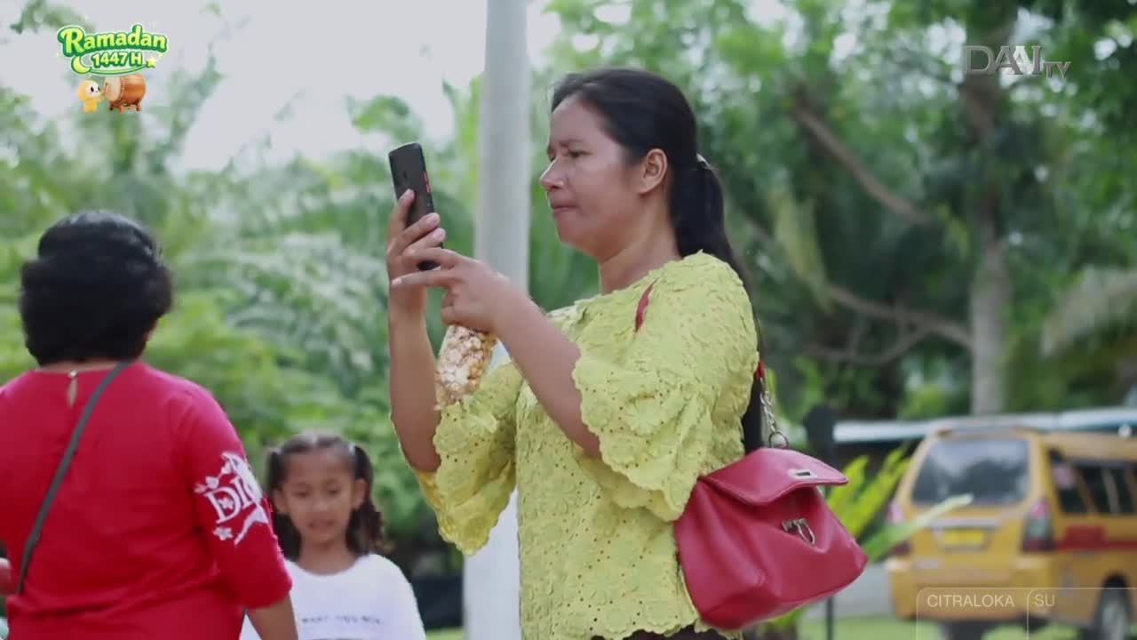 A woman in a bright yellow top holds up her phone, seemingly recording something. A young girl in a white shirt stands behind her, looking towards the camera. A woman in a bright yellow top holds up her phone, seemingly recording something. A young girl in a white shirt stands behind her, looking towards the camera.