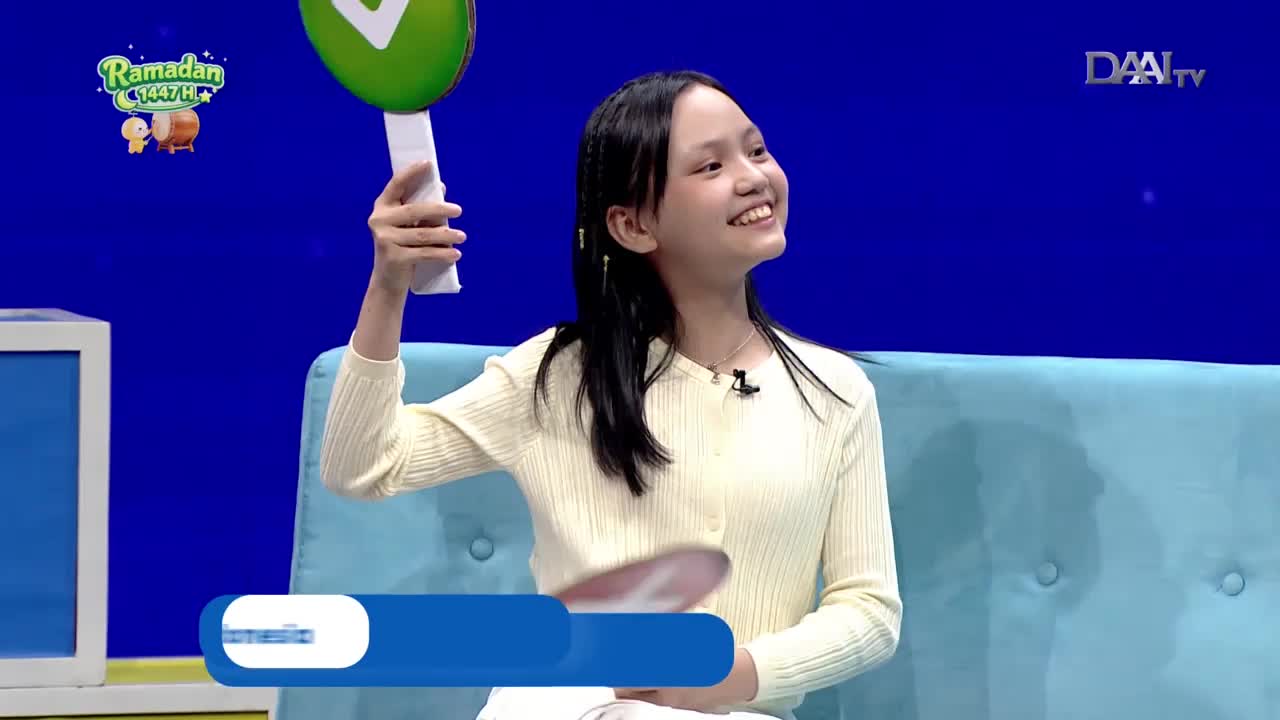 A young girl in a cream-colored cardigan smiles brightly, holding a white paddle with a green circle at the top. She's on a light blue couch, likely during a DAAI TV broadcast in Indonesia.