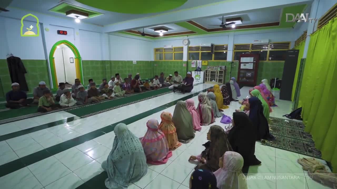 A group of children and adults sit on prayer mats in a room, facing a man reading from a book. Rows of women in colorful hijabs are seated on the floor, their backs to the camera.