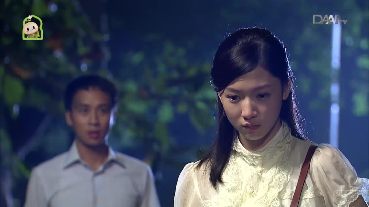 A young woman with dark hair looks down, her expression troubled. Behind her, a man in a light-colored shirt stands with his mouth slightly open, as if speaking.