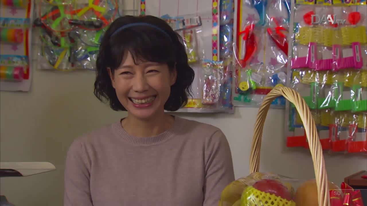 A woman beams, her eyes crinkling at the corners, as she stands behind a basket of fruit. Behind her, colorful toys are neatly arranged on display, the DAAI TV logo subtly visible on the wall.