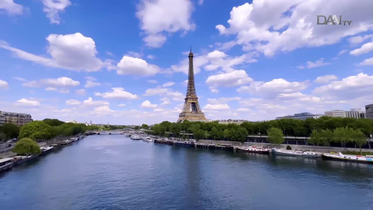 The Eiffel Tower stands tall against a bright blue sky dotted with fluffy white clouds. Barges line the Seine River, their reflections shimmering on the water's surface.
