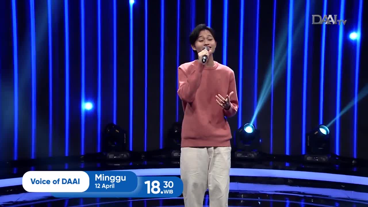 A young man stands center stage, holding a microphone and singing. He's dressed in a coral sweater and light-colored pants, bathed in the blue lights of the "Voice of DAAI" broadcast. A young man stands center stage, holding a microphone and singing. He's dressed in a coral sweater and light-colored pants, bathed in the blue lights of the "Voice of DAAI" broadcast.