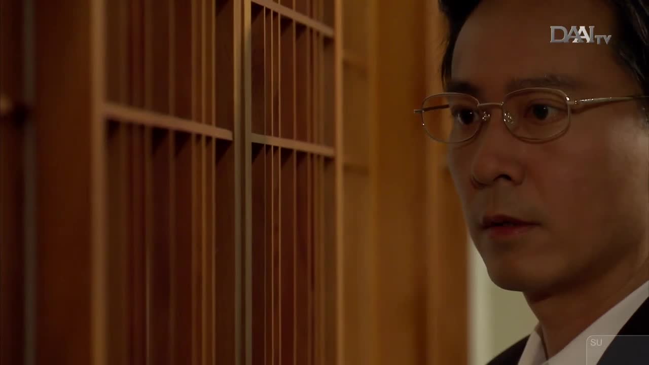 A man in glasses looks intently to his left, his brow furrowed. Behind him, a wooden screen with vertical slats casts subtle shadows. A man in glasses looks intently to his left, his brow furrowed. Behind him, a wooden screen with vertical slats casts subtle shadows.