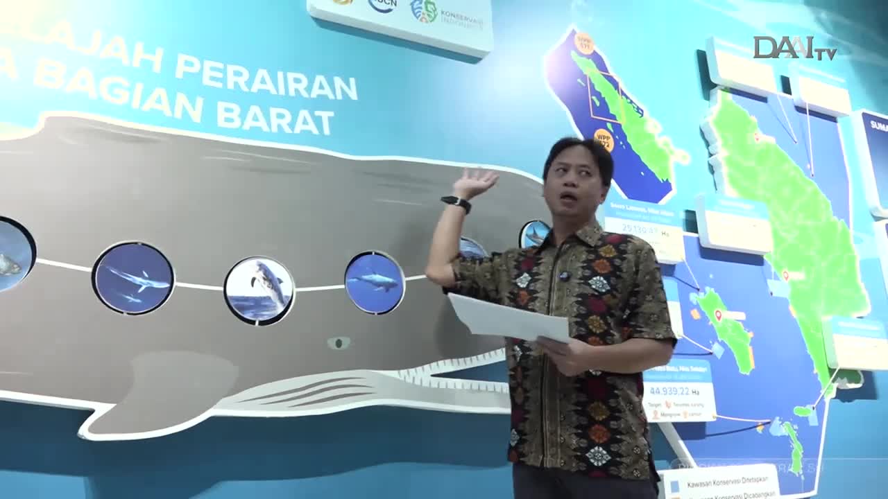 A man in a batik shirt gestures towards a large mural depicting marine life and maps of Indonesia. He holds a paper, likely for a presentation broadcast on DAAI TV.