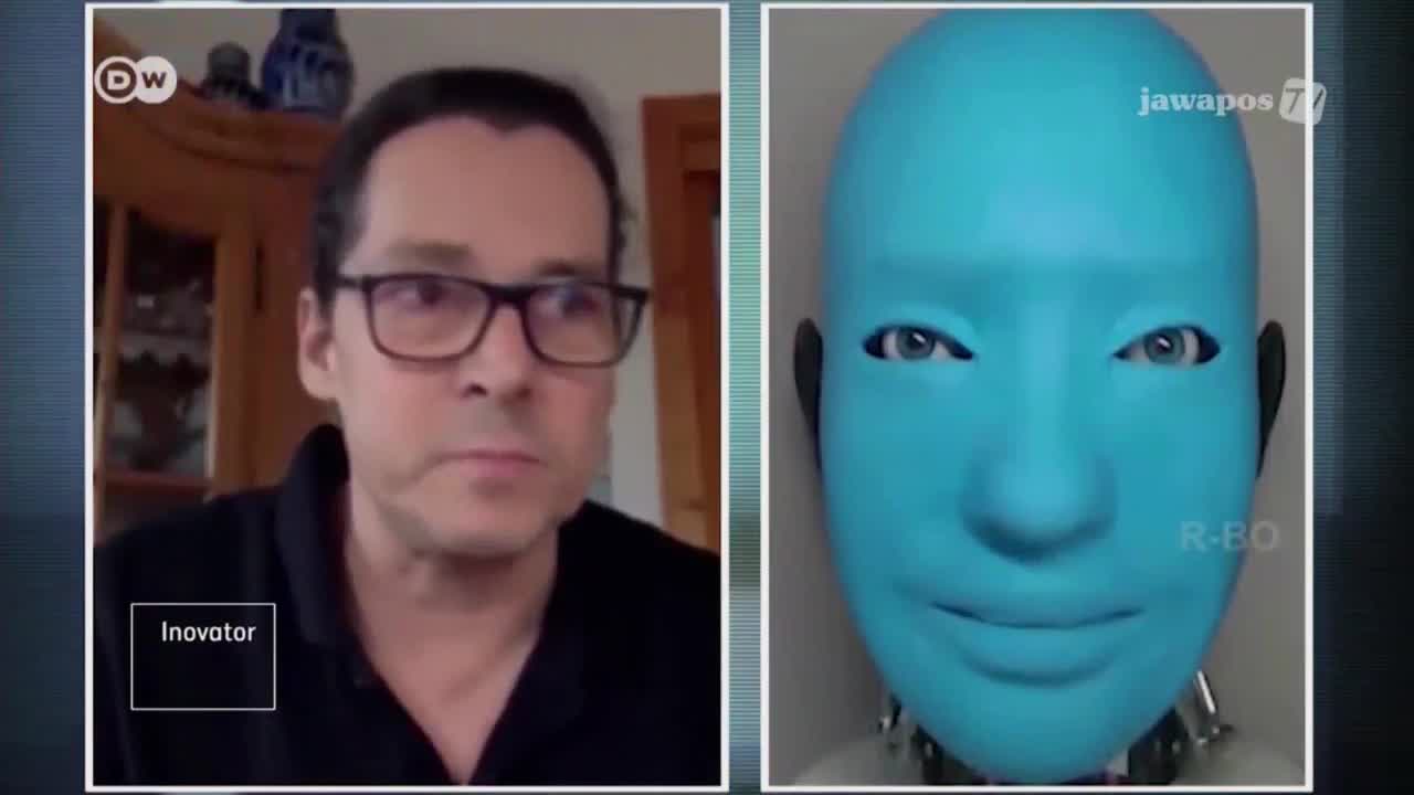 On Jawa Pos TV, a man in glasses is being interviewed, while a blue robot head with realistic eyes sits beside him. The robot's gaze is fixed, and the man seems to be listening intently.
