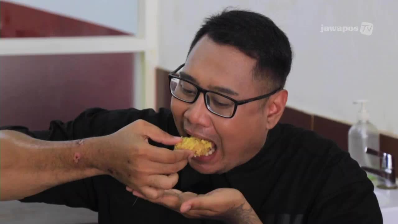 A man, wearing glasses, bites into a piece of fried food, his mouth open wide.  The Jawa Pos TV logo is visible in the corner, as the man enjoys his meal.
