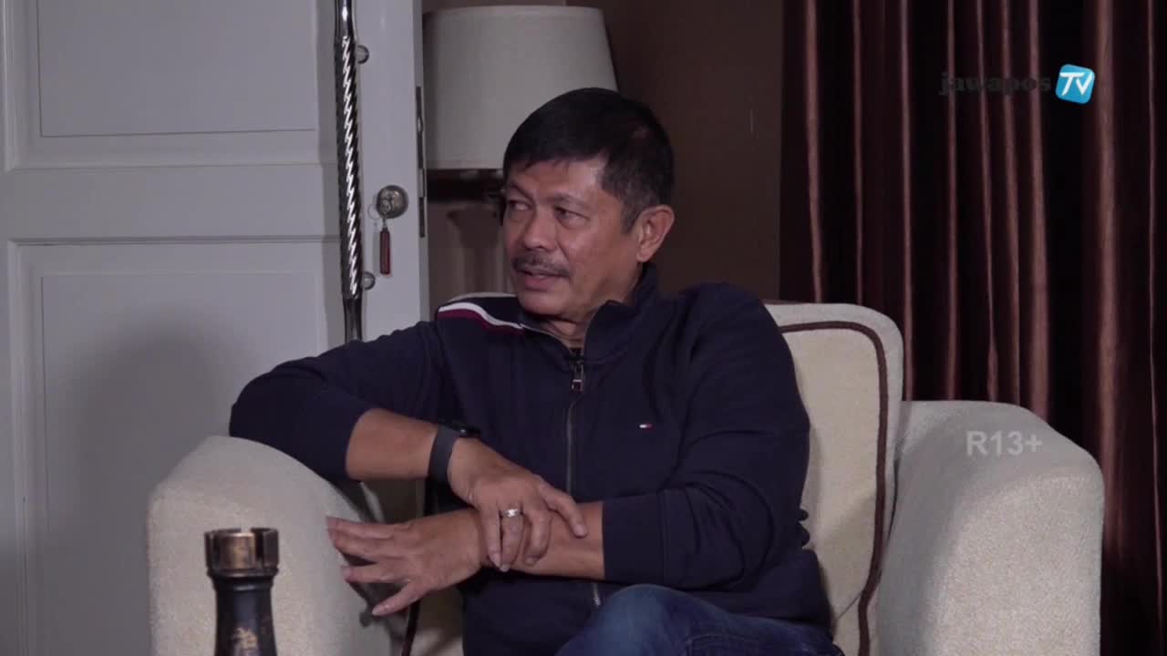 A man in a dark blue jacket sits in an armchair, gesturing with his hands while speaking. The Jawa Pos TV logo is visible in the background.
