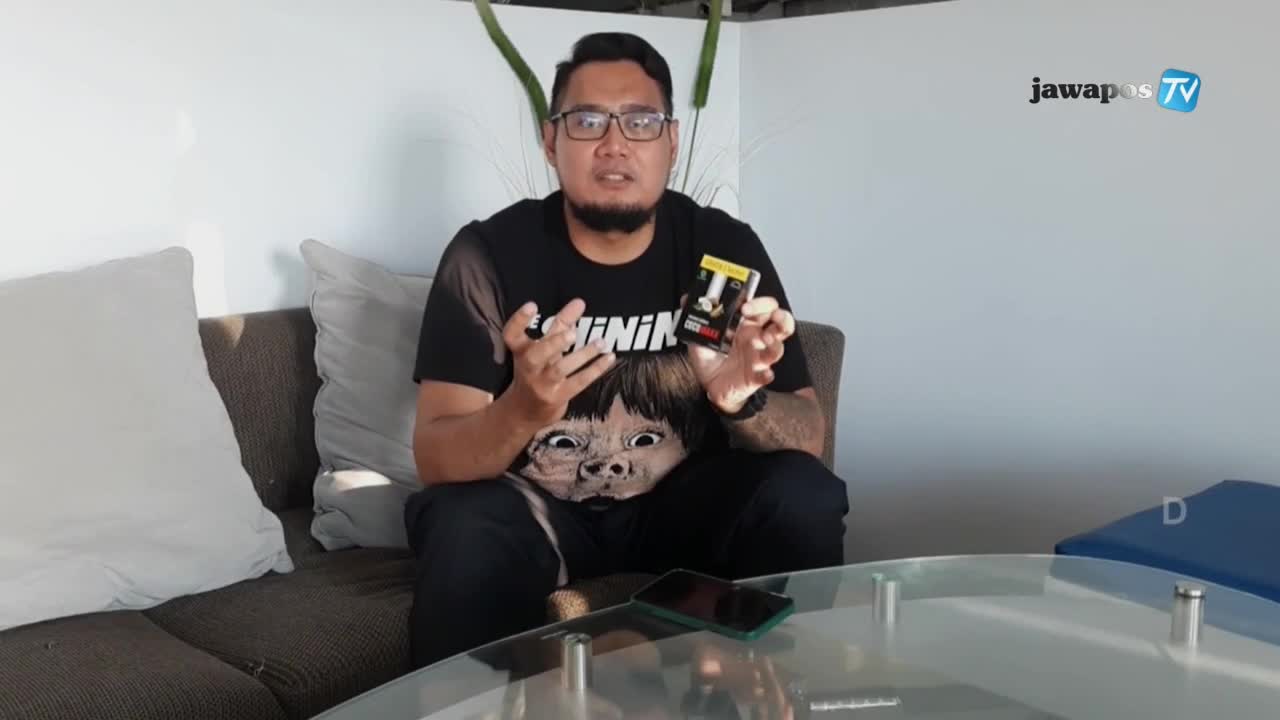 A man in glasses, seated on a couch, gestures with his hands as he holds a small box. The Jawa Pos TV logo is visible in the corner.

