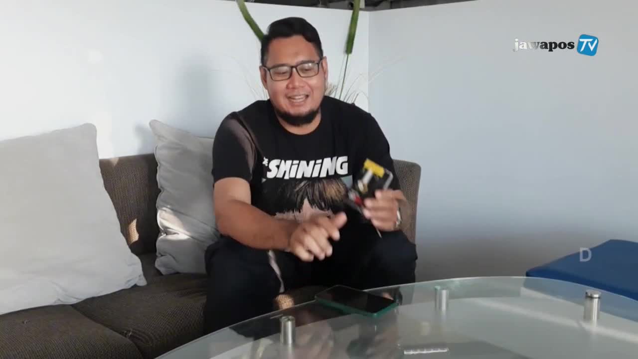 A man in glasses, wearing a "Shining" t-shirt, sits on a couch and gestures with a small device. The Jawa Pos TV logo is in the background.
