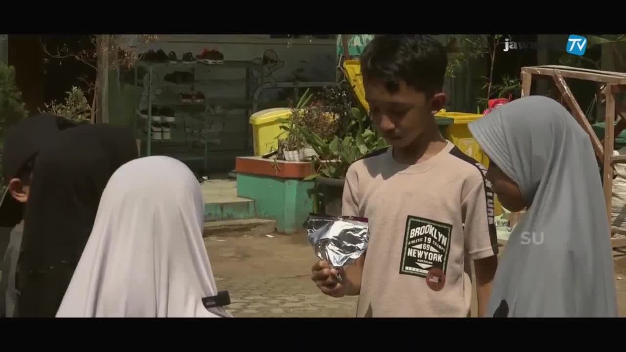A boy holds a small, crinkled foil packet, showing it to two girls. The Jawa Pos TV logo is visible in the corner, suggesting this is a local news report from Indonesia.
