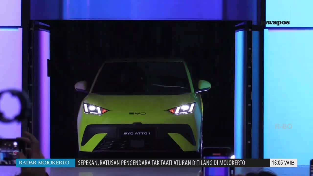 A lime green BYD ATTO 1 electric car is slowly emerging from a dark enclosure, its headlights blazing. The Jawa Pos TV logo is visible, and the news ticker below discusses traffic violations in Mojokerto, Indonesia.
