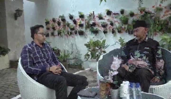 Two men sit facing each other in wicker chairs, framed by a wall of potted plants. The Jawa Pos TV interview is taking place outdoors, under a white wall.
