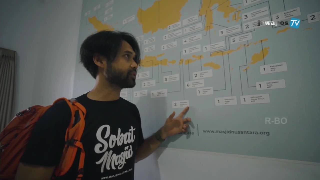 A man, wearing a backpack and a "Sobat Masjid" t-shirt, gestures towards a map of Indonesia on the wall. The Jawa Pos TV logo is visible in the corner.
