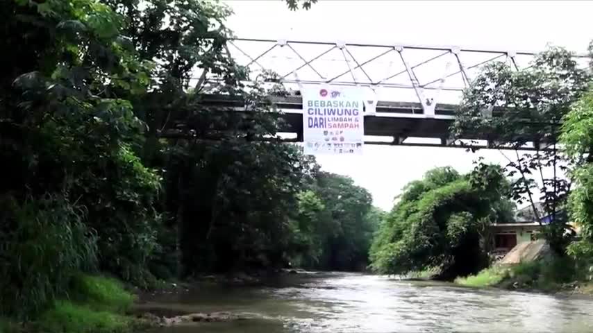 A river flows under a metal bridge, its surface reflecting the overcast sky. A banner hangs from the bridge, likely part of a Jawa Pos TV report about the Ciliwung River in Indonesia.
